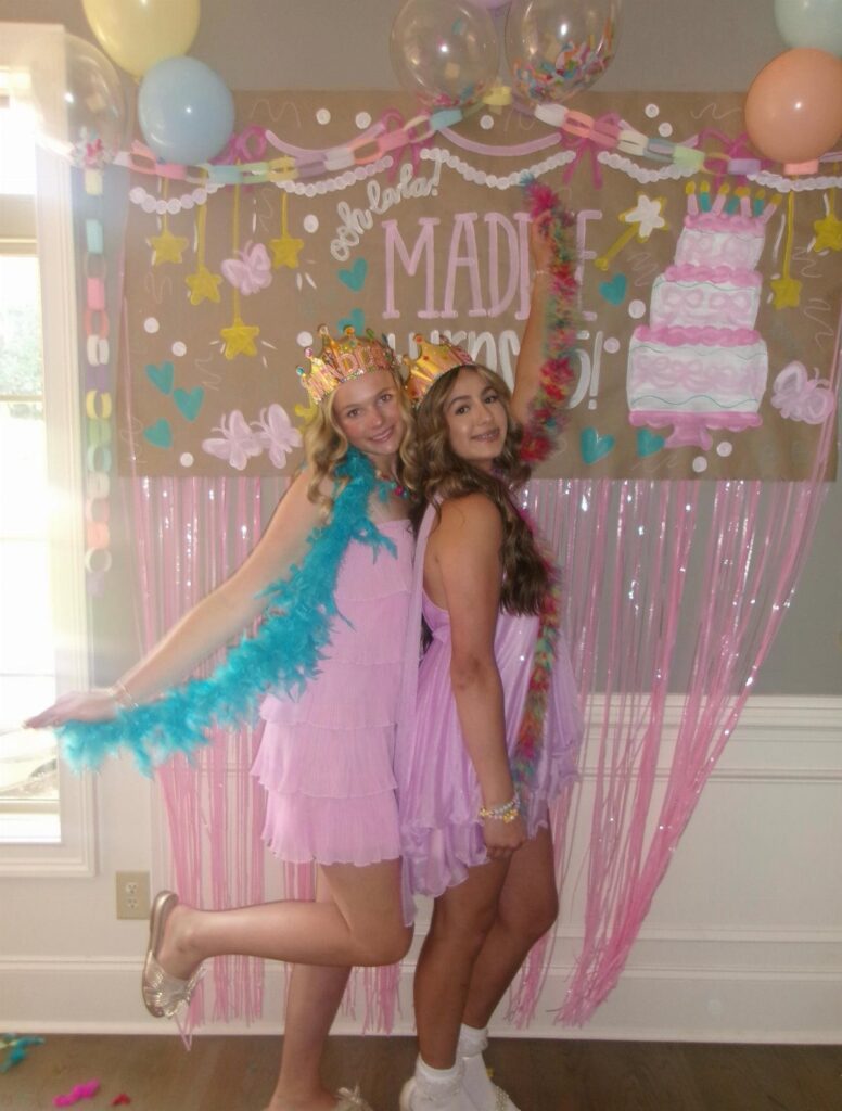 Two teenage girls in pink dresses and crowns posing by a birthday backdrop.