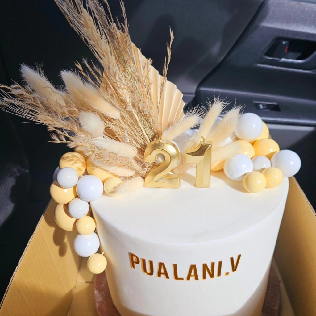 White birthday cake with gold 21 topper, yellow candies, and dried pampas grass.
