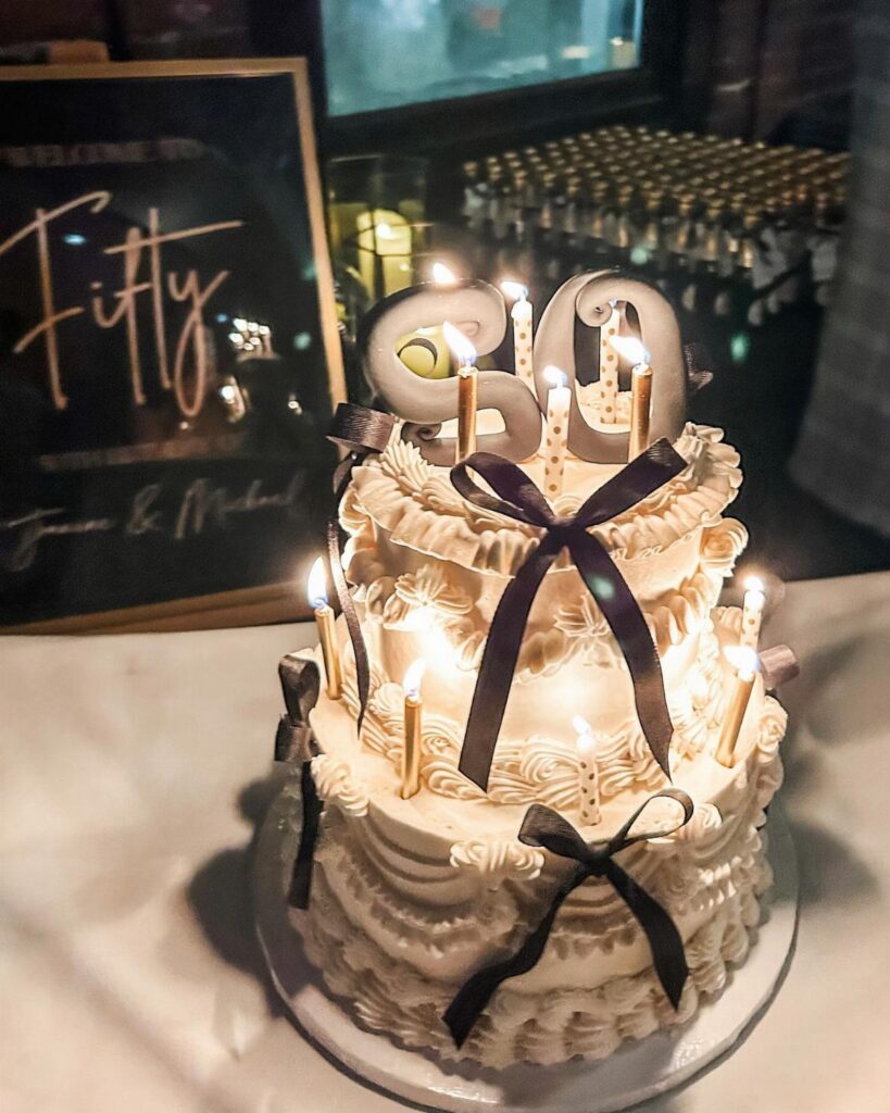 Two-tier white birthday cake with black ribbons and silver number 50 toppers.