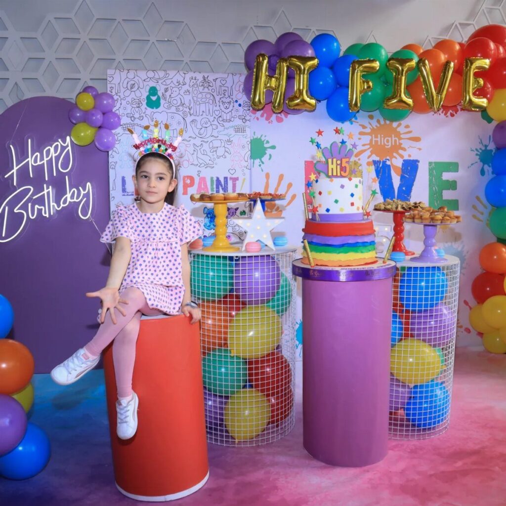 Young girl in a crown sitting amidst colorful balloons and a rainbow cake.
