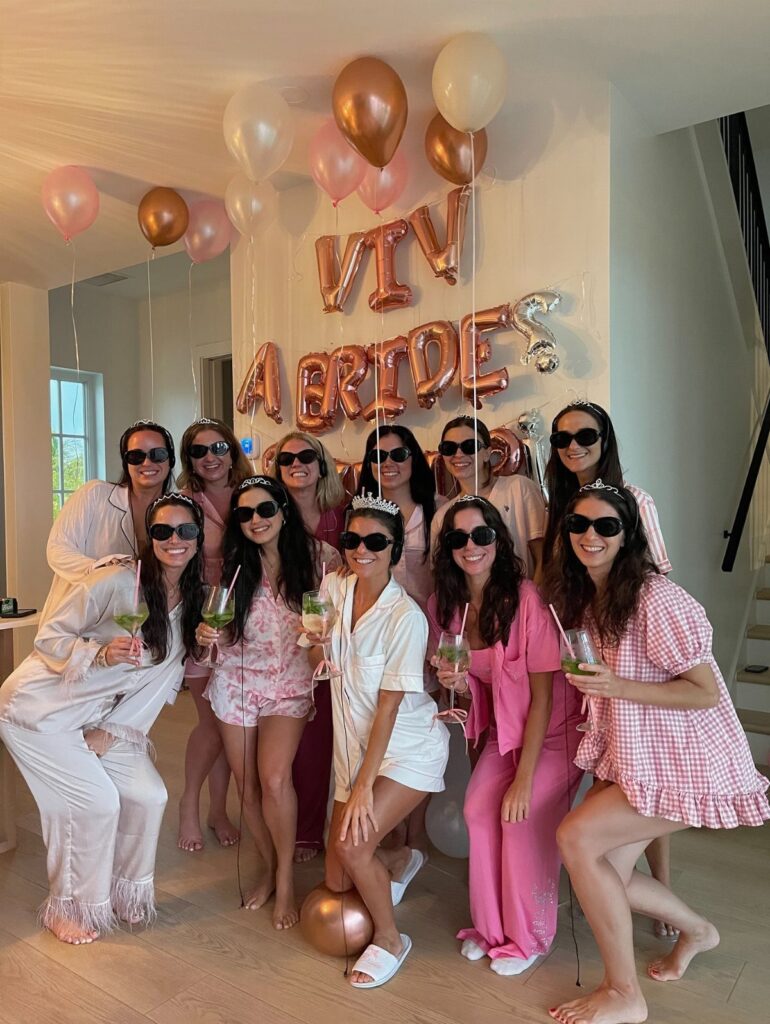 Group of women in pajamas posing with bride-to-be balloons and drinks indoors.