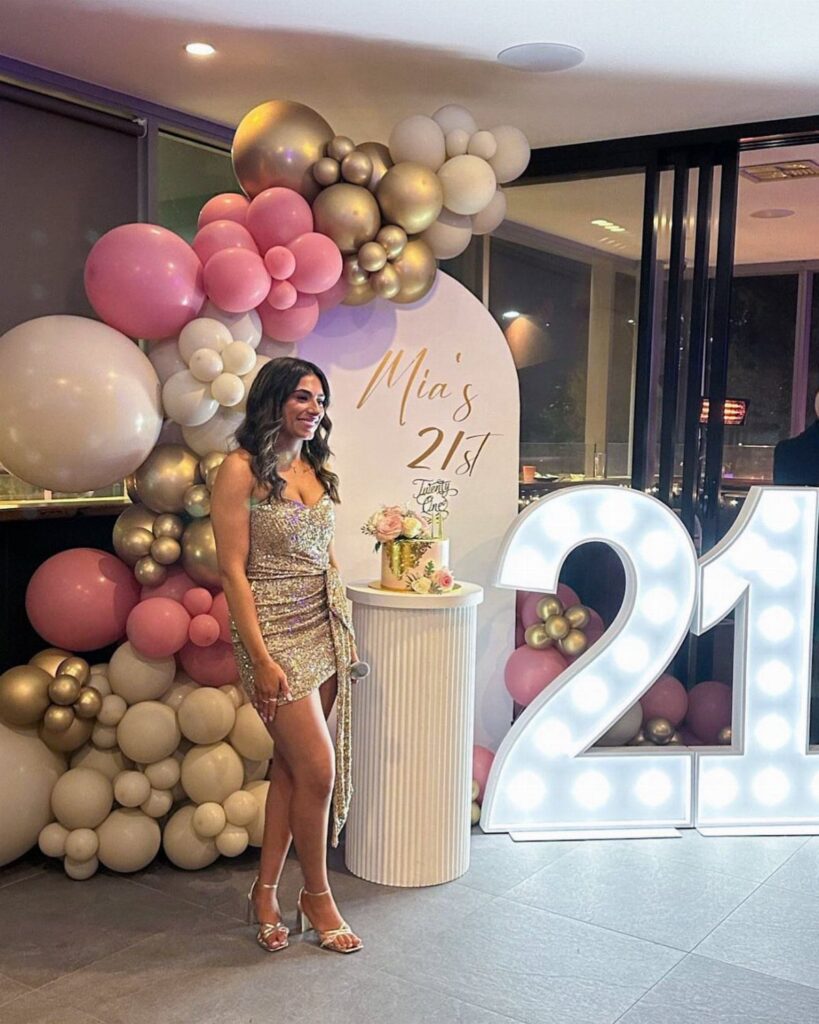 Woman in gold dress posing by 21st birthday balloons and light-up numbers