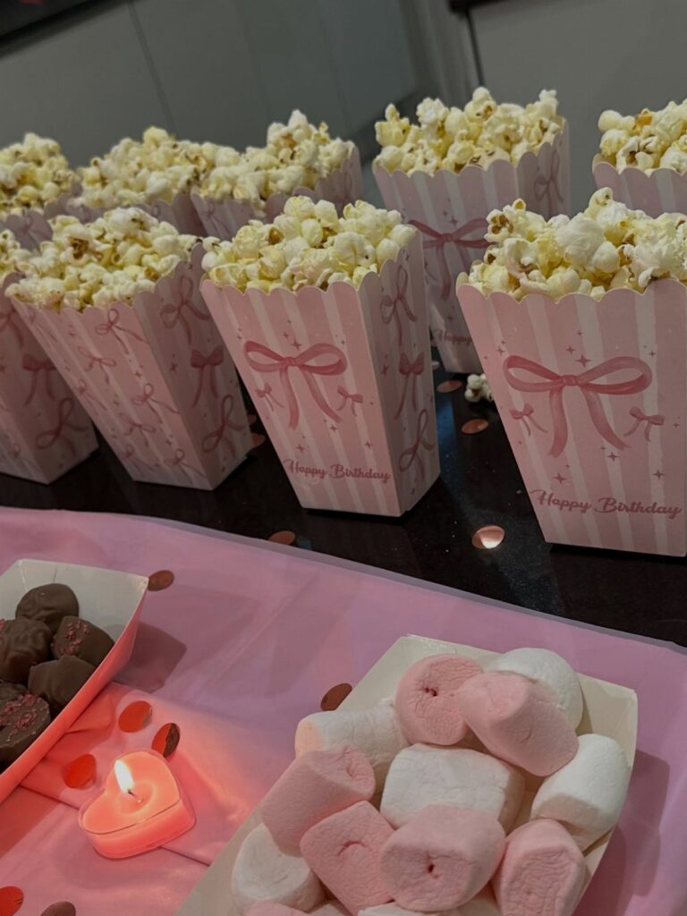Pink birthday popcorn boxes and trays of marshmallows and chocolates on a table.
