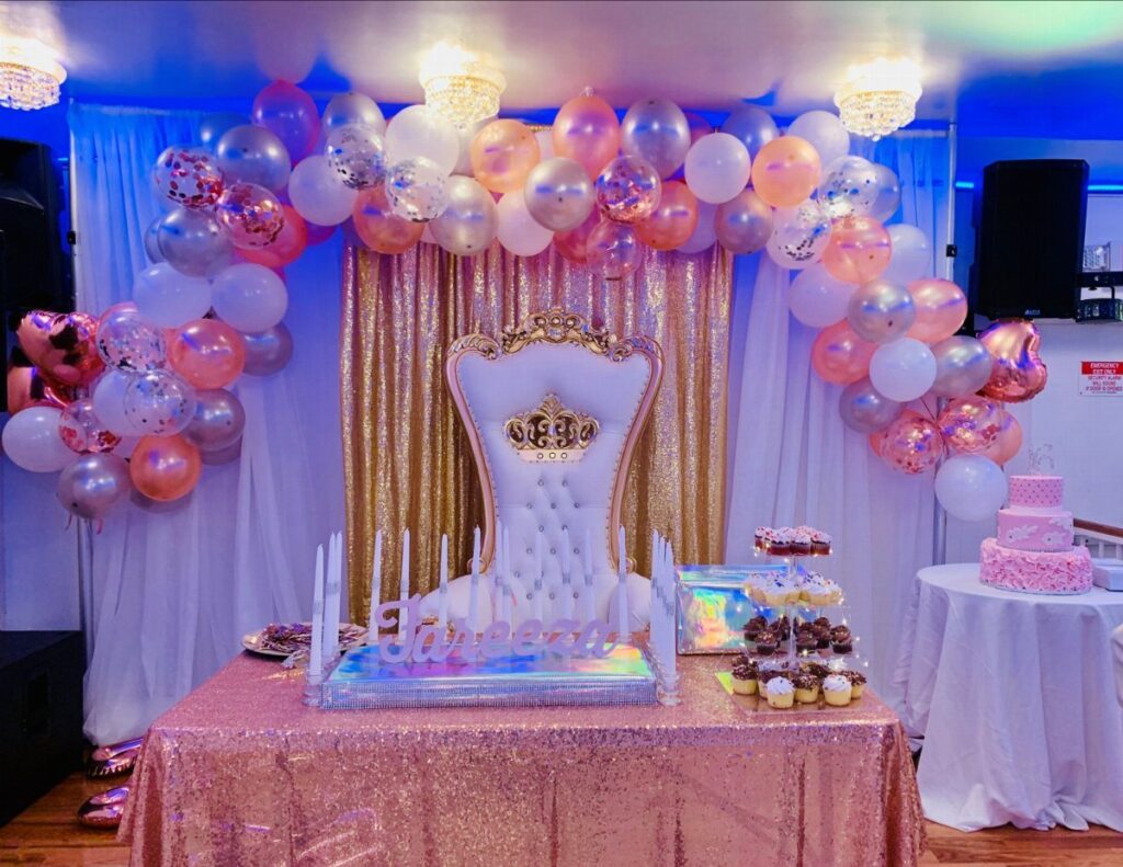 Party setup with rose gold sequin table, white throne, and pink balloon arch.