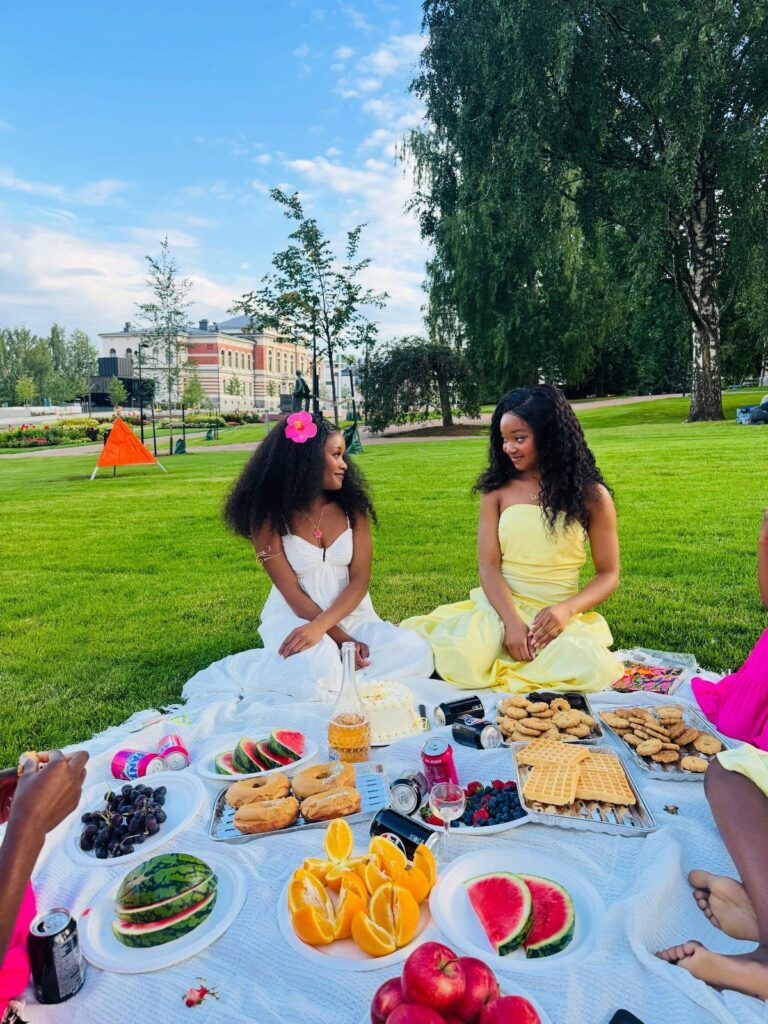 Two women enjoying a picnic spread with fruit and sweets in a sunny park.