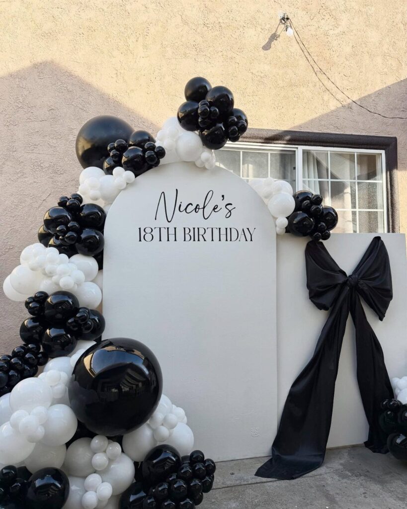 White arched birthday backdrop with black and white balloons and a large black bow.
