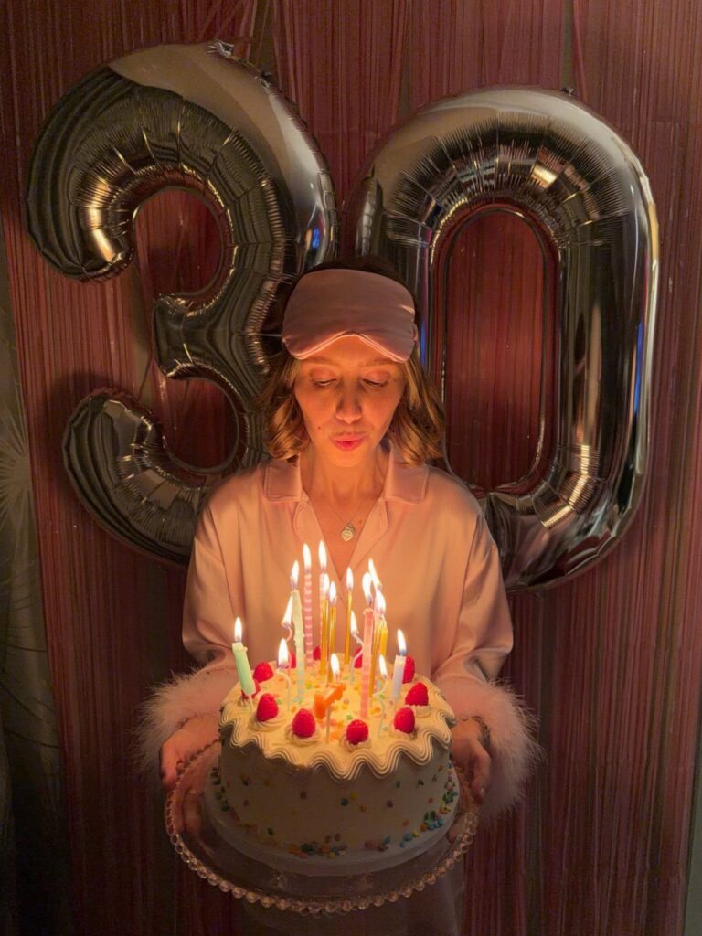 Woman in pink pajamas holding a lit birthday cake before silver 30 balloons