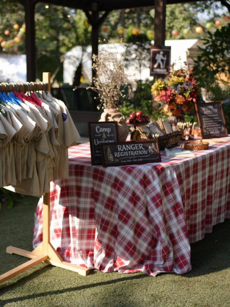 Camping themed party setup with checkered tablecloth, tan shirts, and registration signs.