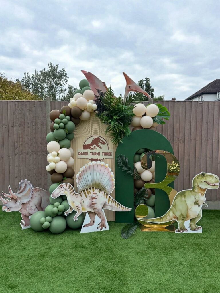 Dinosaur themed birthday party setup with balloons, dinosaur cutouts, and a gold number three.