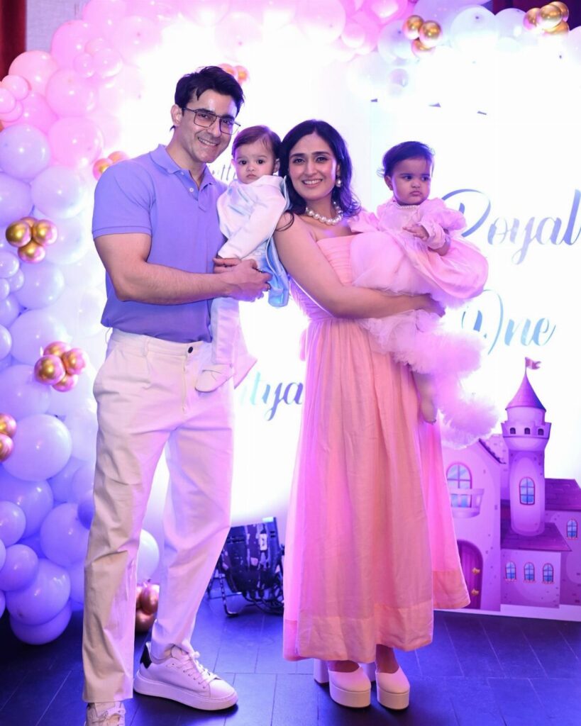 Family posing with toddlers in celebratory white and pink outfits before balloon backdrop.