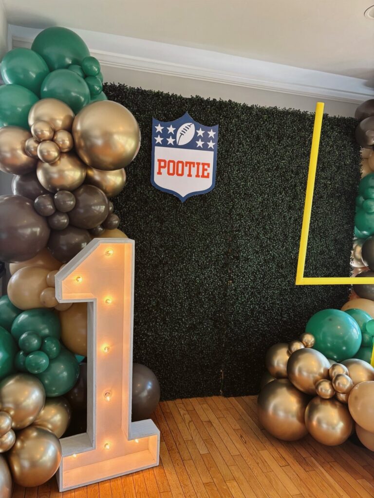 Football themed birthday party setup with green and gold balloons and light-up number one