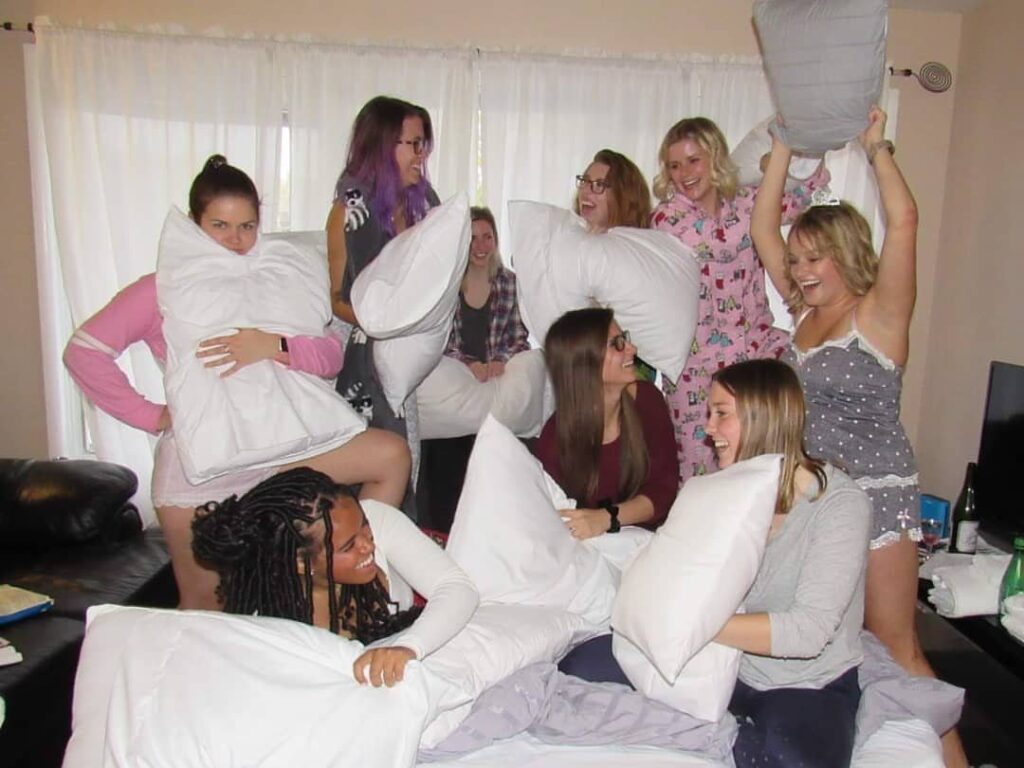 Group of young women laughing and playing with white pillows during a sleepover.