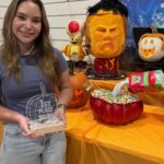 Young woman holding a first place trophy next to carved Halloween pumpkins.