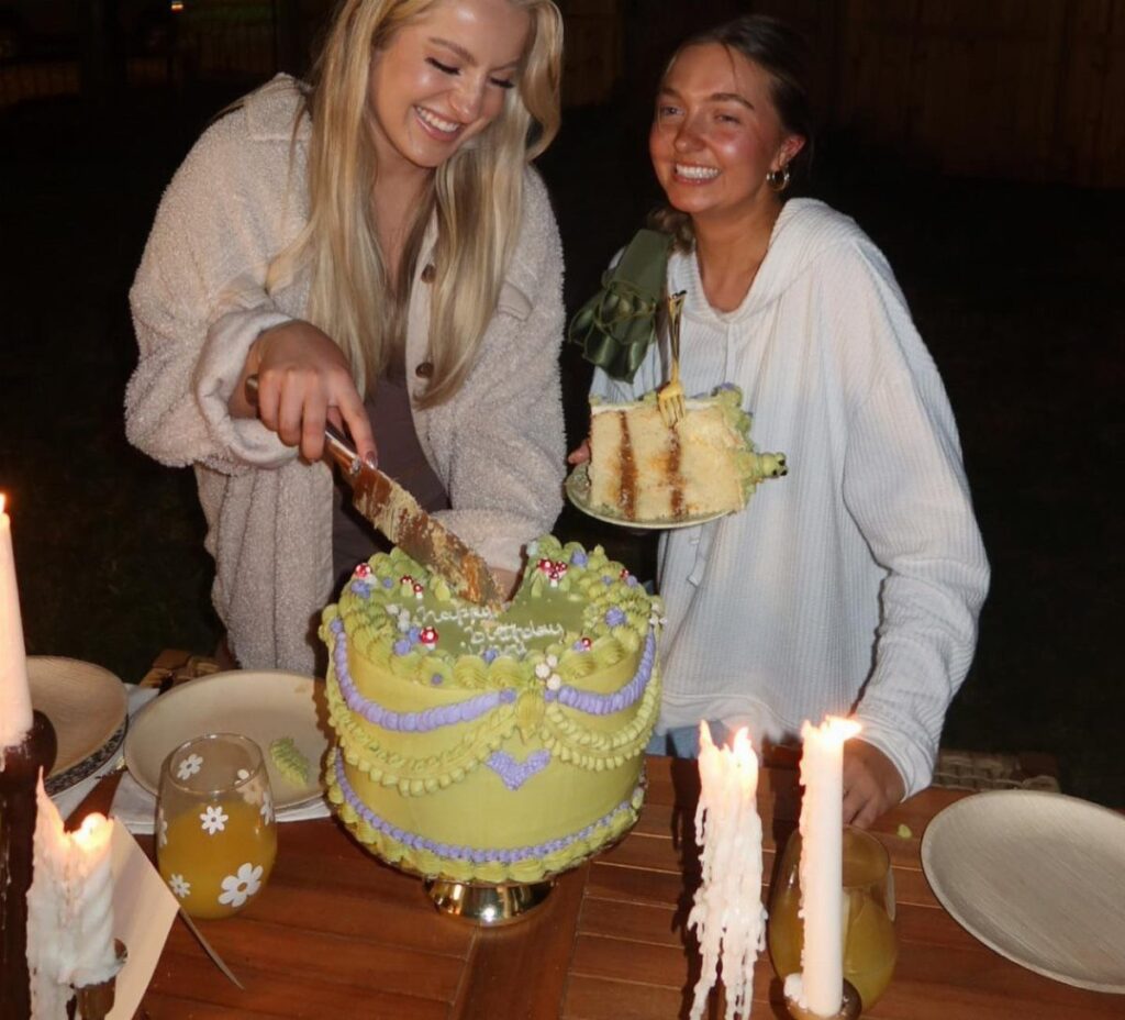 Two women cutting and serving a green birthday cake at an outdoor night party.
