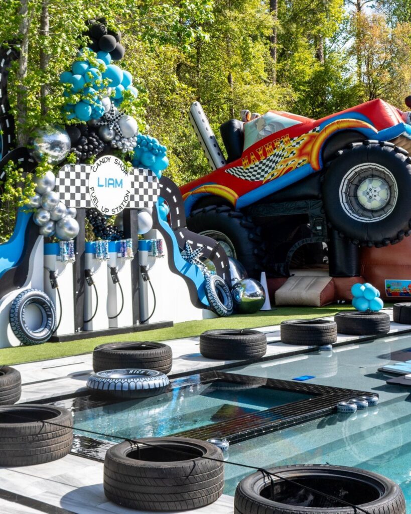Monster truck birthday party setup with inflatable truck, balloons, and tires by a pool.