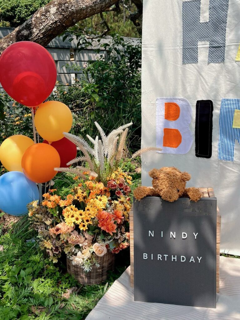 Birthday party decorations with colorful balloons, flowers, and a teddy bear outdoors.