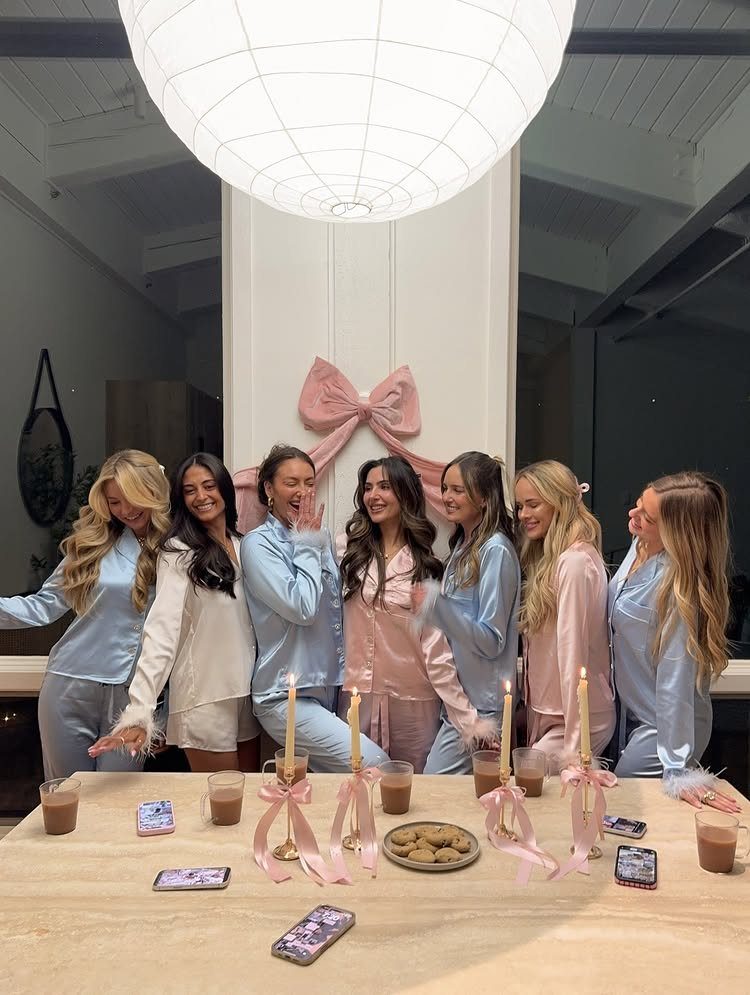 Seven women in satin pajamas posing behind a table with drinks and candles.