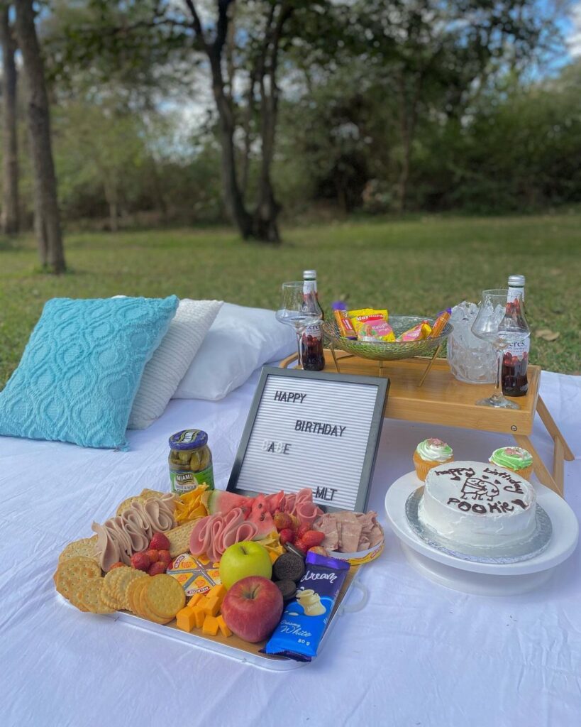 Birthday picnic setup with cake, charcuterie board, and drinks on a white blanket.