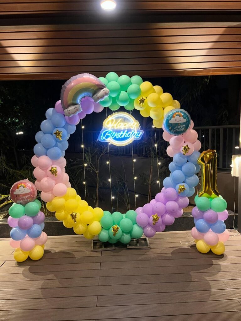 Rainbow balloon arch with a neon Happy Birthday sign on a wooden deck.
