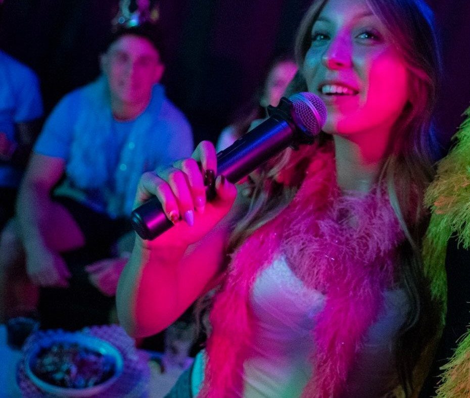 Woman singing into a microphone wearing a pink feather boa at a party.