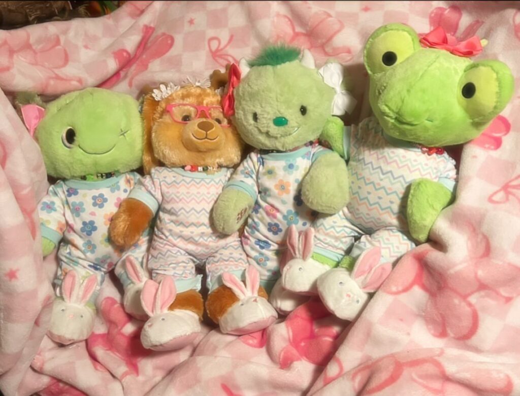 Four plush stuffed animals wearing pajamas and slippers on a pink patterned blanket.