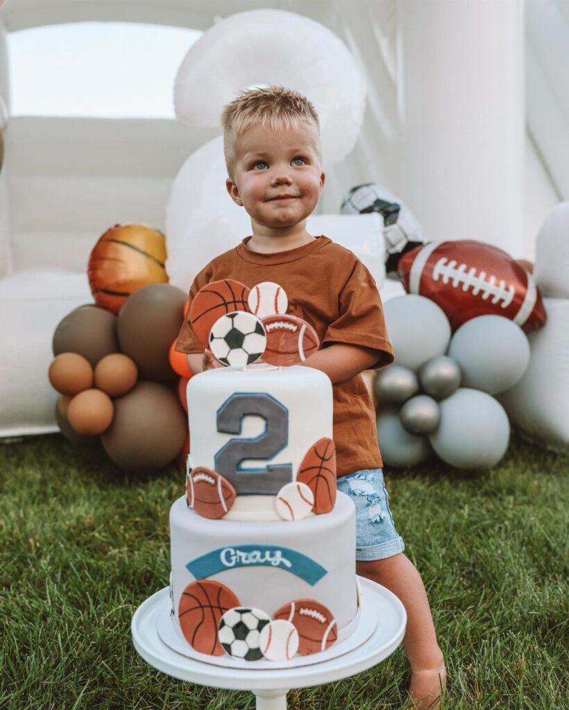 Young boy standing behind a sports-themed birthday cake with balloons and bouncy castle.
