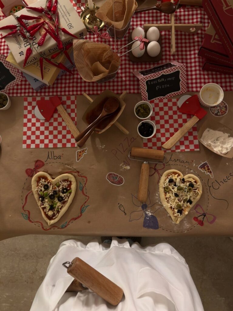 Two heart-shaped pizzas with toppings on a table with rolling pins and ingredients.